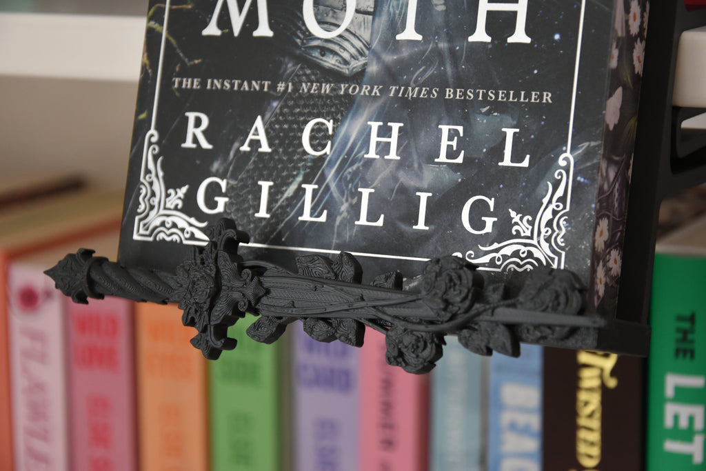 A close-up of a black ornate trim piece shaped like a decorative dagger with roses, holding a book on a shelf with other colorful books blurred in the background.