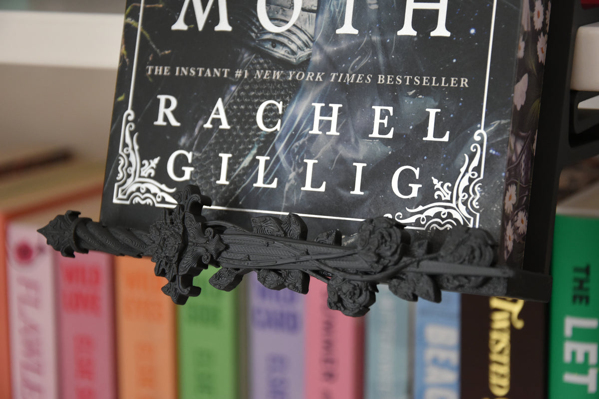 A close-up of a black ornate trim piece shaped like a decorative dagger with roses, holding a book on a shelf with other colorful books blurred in the background.