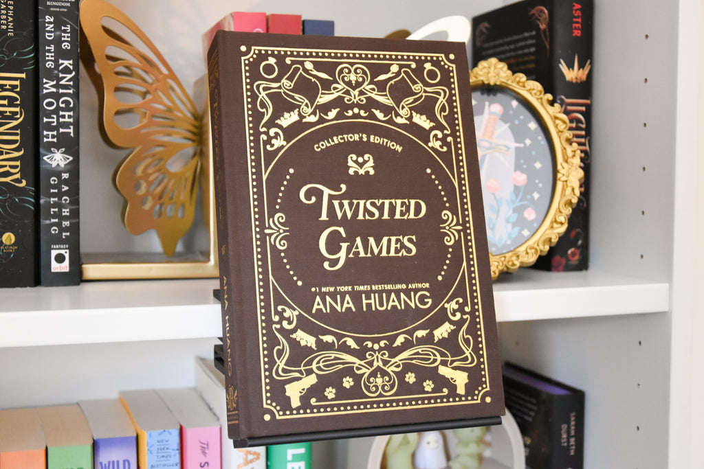 Twisted Games Collector's Edition book by Ana Huang, leaning on a white bookshelf next to other books and decorative items. It is displayed using a black version of a book holder. The book display clamps onto the bookcase shelf. 
