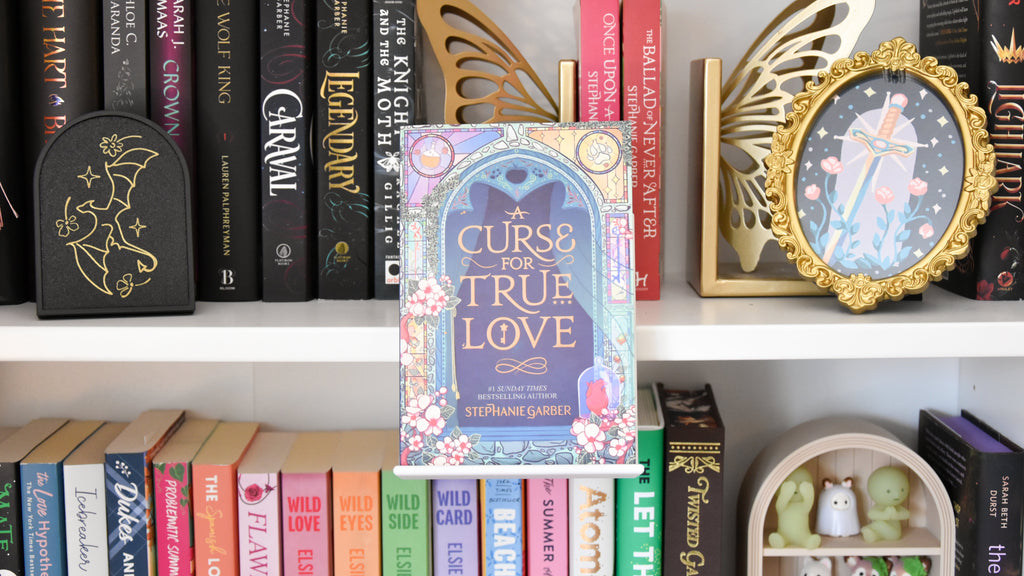 The book 'Curse for True Love' by Stephanie Garber stands upright on a white bookshelf, surrounded by other books, decorative gold angel wings. It is held up by a white book display which clamps onto the bookshelf to create a floating effect. 