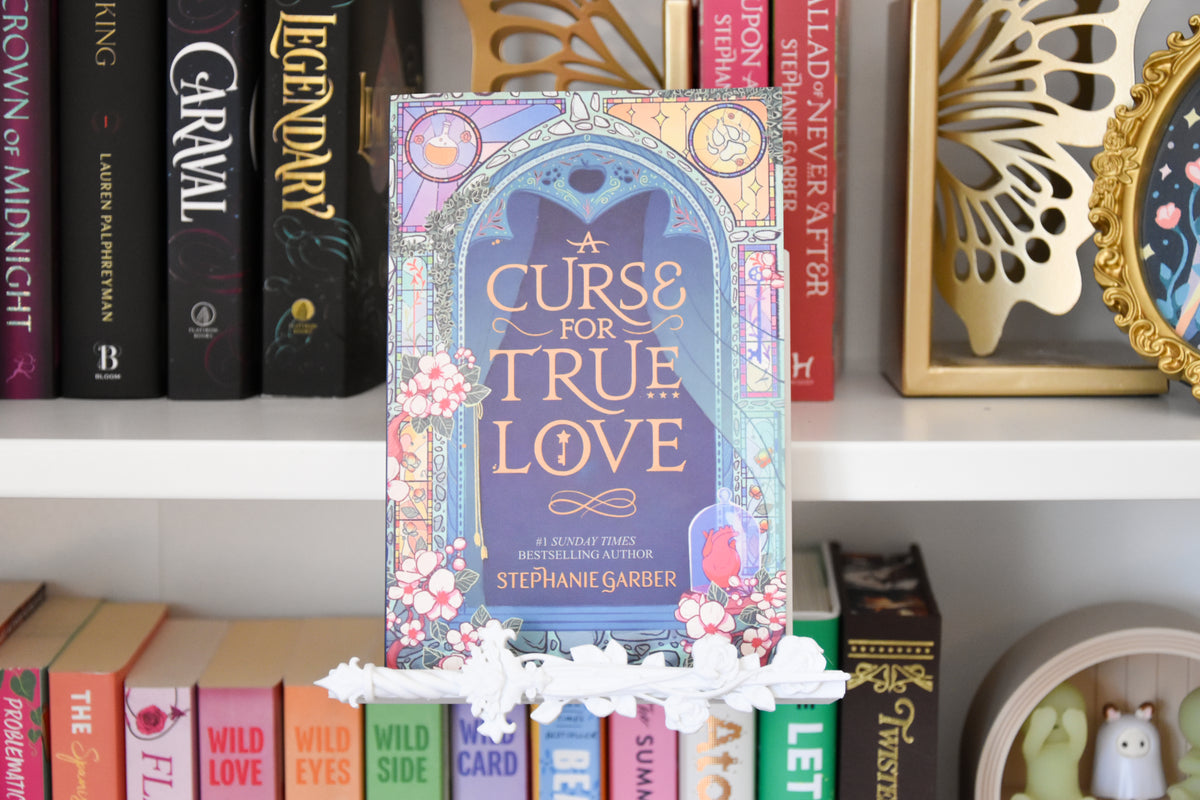 The book 'A Curse for True Love' by Stephanie Garber stands upright on a white bookshelf, surrounded by other books. The cover features a purple background with a cat illustration and decorative pink floral borders. The bottom of the book display features a rose dagger trim piece.