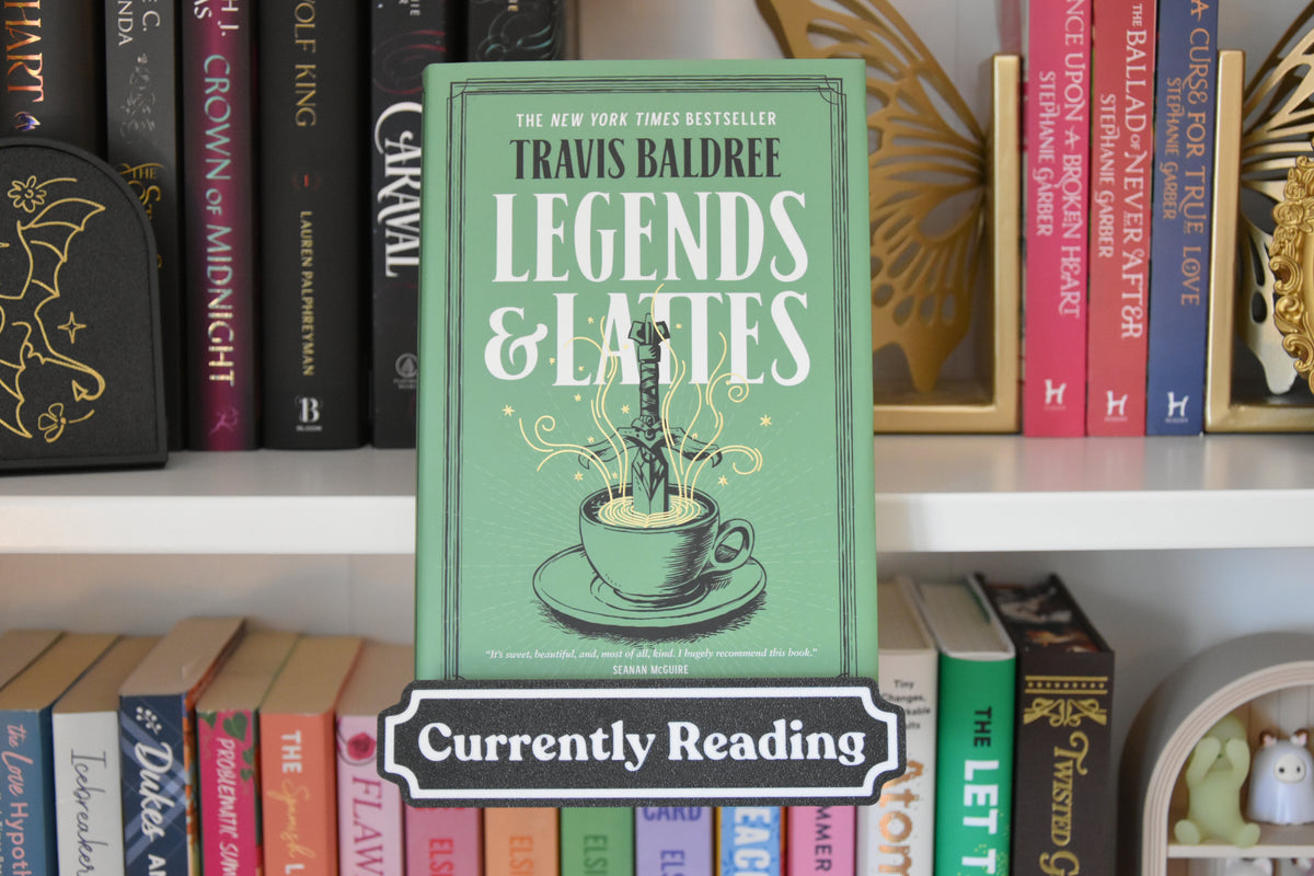 A green book cover for Travis Baldree's 'Legends & Lattes' stands upright on a white bookshelf, surrounded by other books. A white 'Currently Reading' label is overlaid at the bottom.
