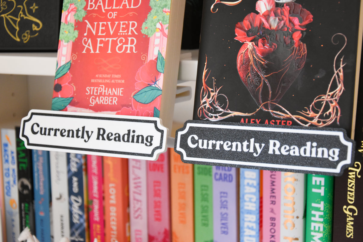 Two decorative 'Currently Reading' bookshelf signs displayed in front of two different books on a shelf.
