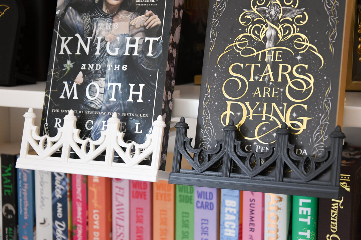 Two books displayed on a black decorative book stand, with a white decorative book stand holding 'Knight and the Moth'. The black stand holds a book with gold lettering that reads 'The Stars Are Dying'. Both stands feature trim inspired by gothic architecture.