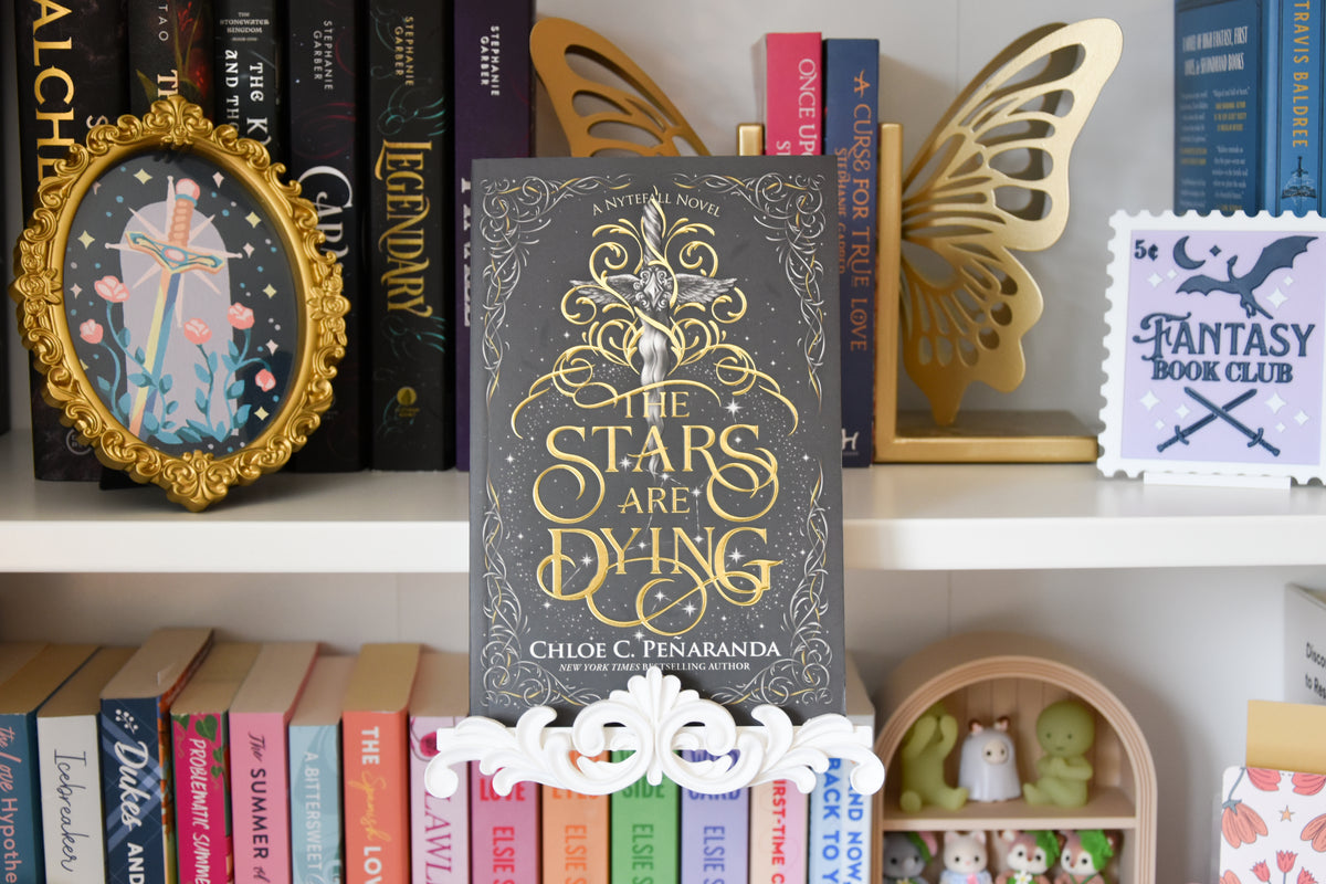 The Stars Are Dying book by Chloe C. Penaranda displayed on a white shelf. The book is displayed using a white book stand that clamps onto the bookshelf. The stand features a decorative flourish trim piece at the bottom.