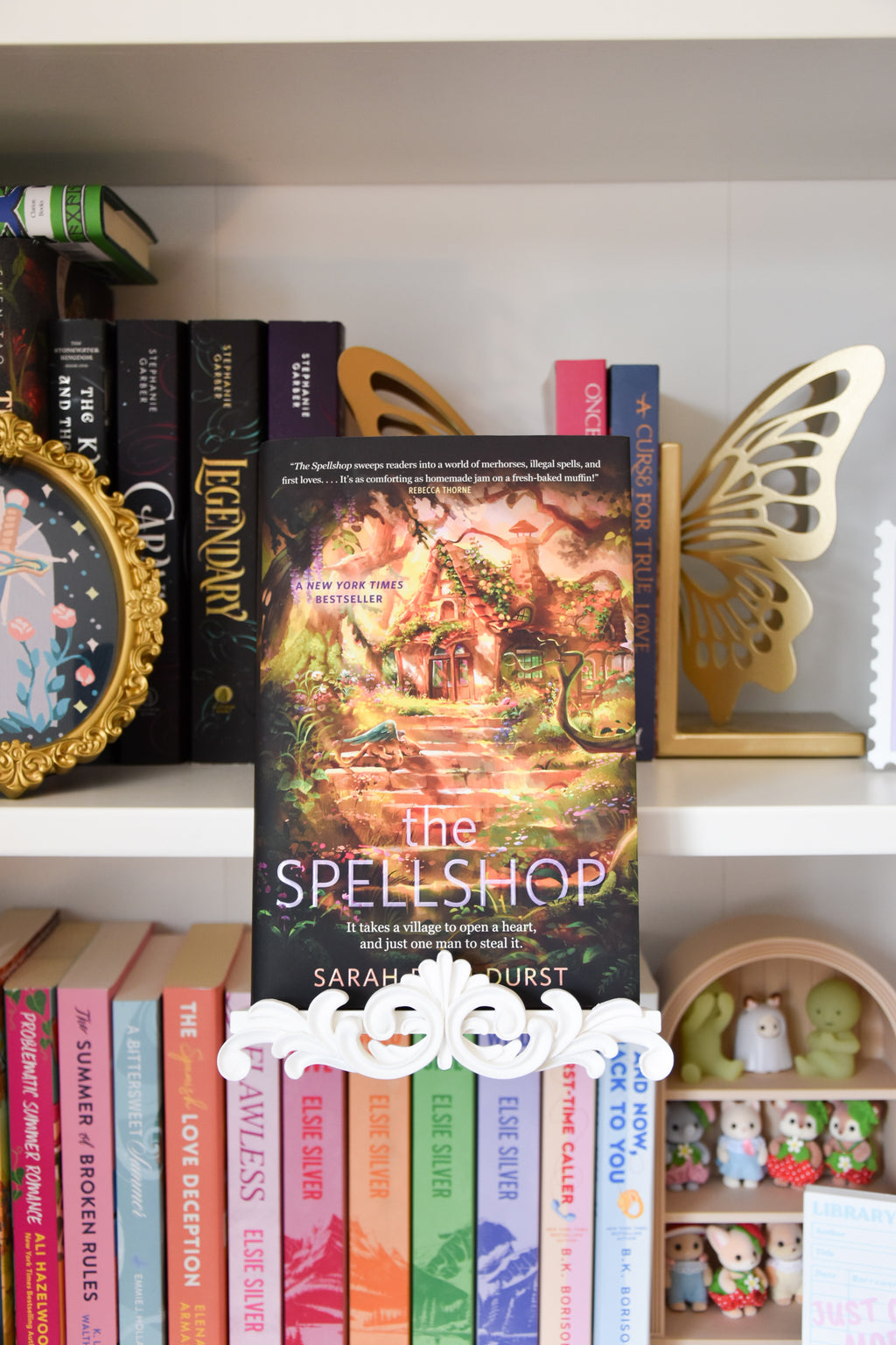 The Spellshop book is displayed using a white book stand that clamps onto the bookshelf. The stand features a decorative flourish trim piece at the bottom.