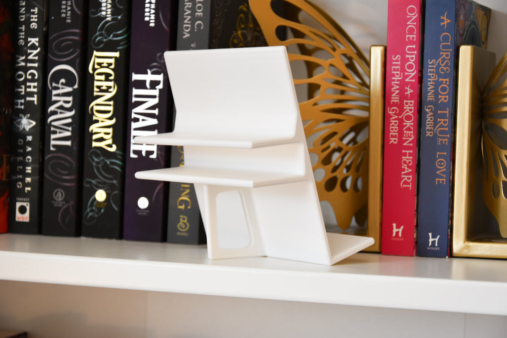 A white modern book display is shown from the back. It features a clamp for a bookshelf, and a fin that acts as an easel. 