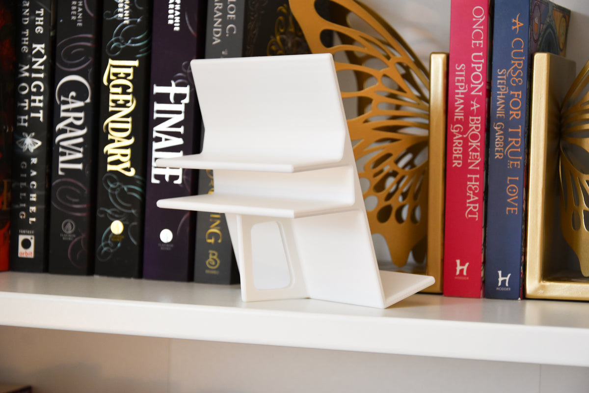 A white modern book display is shown from the back. It features a clamp for a bookshelf, and a fin that acts as an easel. 